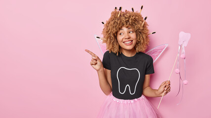 Obraz na płótnie Canvas Photo of good looking cheerful woman dressed like tooth fairy indicates index finger aside on blank space demonstrates place for your advertisement isolated over pink background. Tooth care concept
