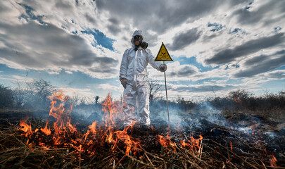 Firefighter ecologist fighting fire in field. Man in protective suit and gas mask near burning grass with smoke, holding warning sign with skull and crossbones. Natural disaster concept.
