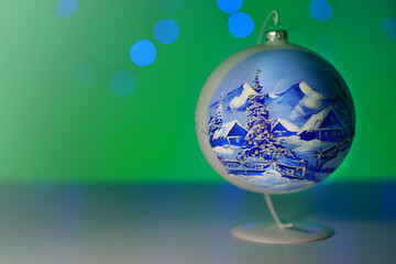 Fototapeta na wymiar Christmas bauble with a winter mountain landscape (hut, snow, Christmas trees, mountains) standing on a stand. Picture with blue blurred Christmas tree lights. Green background. Merry Christmas. 