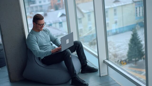 Comfortable work conditions in the modern office. Happy young man sitting in bean bag chair typing on his laptop at looking at window. High angle view.