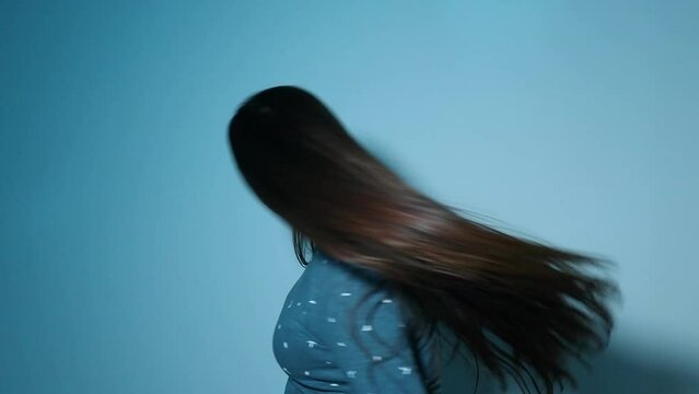 White woman whipping her long brown hair while spinning herself around in a blue room and smiling in the end