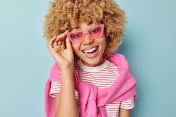 Happy young woman with curly hair and toothy smile keeps hand on pink trendy sunglasses hears good news dresed casually isolated over blue background. Positive human emotions and style concept