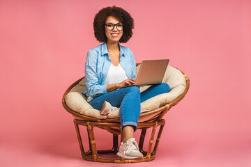 Young african american positive cool lady with curly hair using laptop and smiling isolated over pink background.