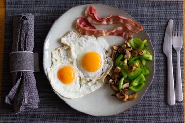 Top view of two fried eggs with bacon, green peppers and mushrooms. Traditional English breakfast with napkin and cutlery, ketogenic diet concept
