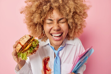 Positive overjoyed female student has lunch break at university holds delicious burger and folders...