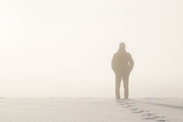 Young adult man silhouette standing in nature mist and looking far away. Thinking about life. Back view. Fresh human boots footprints in fresh deep snow. Cold snowy winter day. Empty place for text. - obrazy, fototapety, plakaty