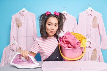 Serious housewife with two pony tails holds basket of laundry for ironing dressed in striped t...