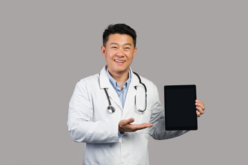 Happy mature chinese man therapist in white coat with stethoscope shows hand on tablet with empty screen