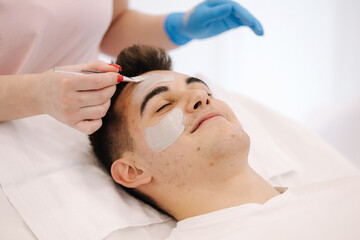 Fototapeta na wymiar Cosmetologist applying clay mask on man's face in spa salon. Close-up of male face during procedure. Pimples and blackheads on the skin. Acne