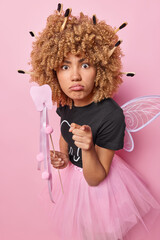Indoor shot of serious attentive woman points index finger directly at camera holds tooth shaped wand reproaches you for doing something bad dressed like fairy isolated over pink background.