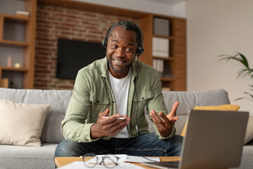 Glad middle aged black man in headphones has video call on computer, supports customers in living room