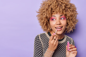 Photo of good looking young woman with curly hair applies lip gloss wears bright makeup undergoes beauty procedures before dating isolated over purple background blank space for your promotion