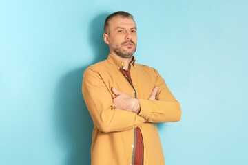 People emotions concept. Portrait of serious mature man posing with folded arms, standing on blue studio background