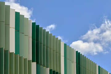 Vertical metal panels of a buildings facade against a blue sky with white clouds. Panels in shades of green. Frame divided diagonally between the building's exterior and the sky