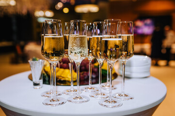 Glass wine glasses with champagne stand on a round white table with fruits and dishes in a restaurant. Photography, banquet, wedding.