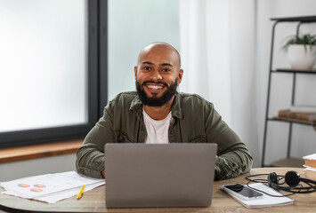 Portrait of happy latin male freelancer sitting at table, working on laptop at home office interior, copy space