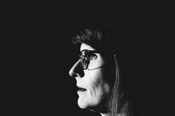Black and white portrait of woman 50plus model with long hair and glasses against black background, dressed in patterned blouse, looking to the side