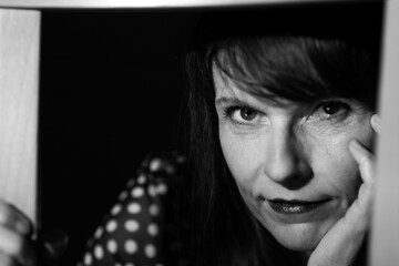 Black and white portrait of woman, model 50plus, in front of black background, sitting behind chair...