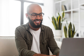 Happy latin businessman working on laptop computer, wearing earbuds and looking at screen, sitting at home interior