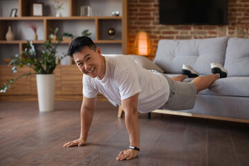 Strong middle aged asian man doing push ups with legs on sofa, working out at home and smiling at camera