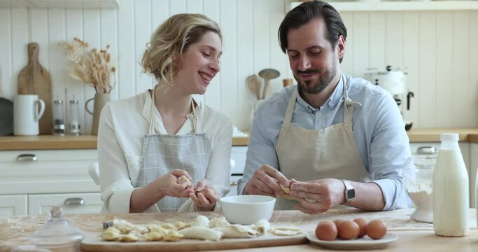 Beautiful millennial couple in aprons sit at table, talking, preparing dough for home-made pastries in modern kitchen at home, enjoy conversation spend weekend, cooking together. Relations, culinary