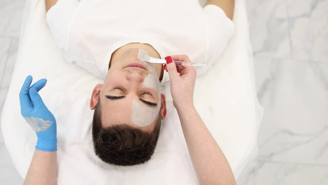 Cosmetologist applying clay mask on man's face in spa salon. Beautician make beuty for handsome man in beauty studio