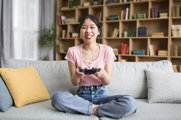 Happy korean lady with joystick, sitting on couch and playing online games in living room interior, copy space