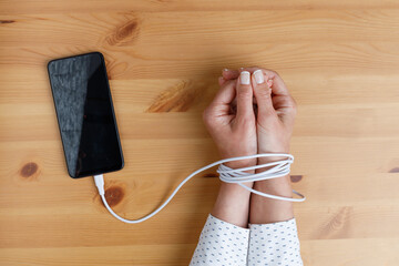 Woman's hands wrapped on wrists with mobile phone cable as handcuffs. Addiction to social networks - obrazy, fototapety, plakaty