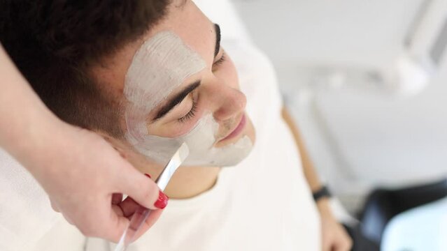 Cosmetologist applying clay mask on man's face in spa salon. Beautician make beuty for handsome man in beauty studio. Vertical video