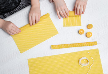Two kids make natural rolled beeswax candles from pressed beeswax honeycomb sheet at home indoors....