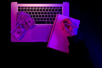 Laptop, blank notepad and paper money in neon lighting, top view.