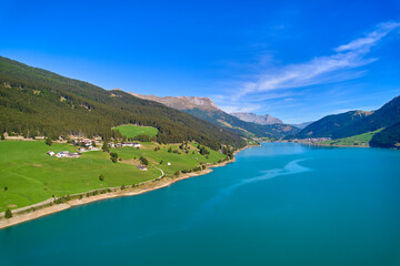 Fototapeta premium Aerial view of lake (Reschensee). Large reservoir surrounded by mountains at sunny noon. Recreation area for tourists and sportsmen. Organic farm on the shore. Italy, Vinschgau, Giern.