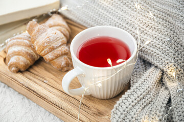 A cup of red tea, a croissant and a knitted element on a wooden tray.