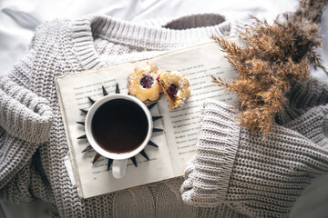 Home composition with a cup of coffee, cookies, a book and a knitted sweater.