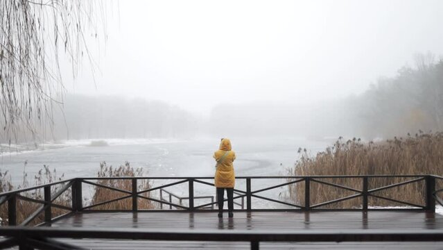 Young man in yellow clothes standing alone on wooden footbridge and staring at lake in foggy forest. Hooded guy. Back view.