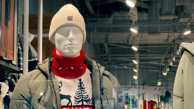 Male mannequin in winter clothes: a reindeer sweater, a down jacket and a knitted hat stands in the window of a clothing store