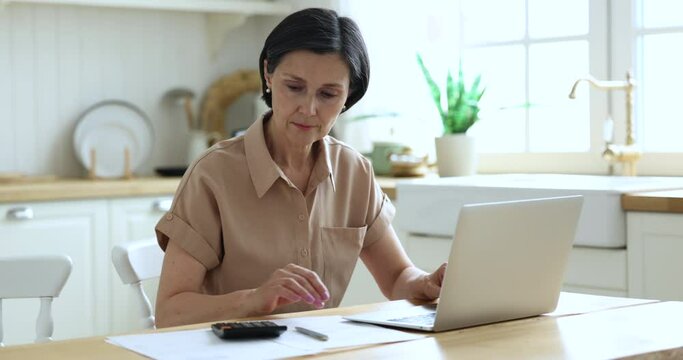 Elderly female sit in home kitchen doing accounting work from home, calculates company financial statement, use calculator, fills data results use laptop app, housewife manage budget, pay bills online