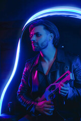 Handsome man in cyberpunk style with a machine gun in his hands in the dark. A guy in a leather jacket in the scenery of the future in neon color. Spaceship.