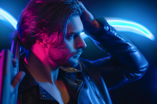 Handsome man in cyberpunk style with a machine gun in his hands in the dark. A guy in a leather jacket in the scenery of the future in neon color. Spaceship.