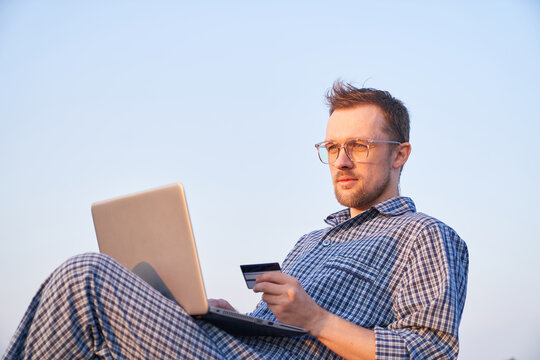 Online shopping concept. Cheerful bearded caucasian male in eyeglasses making shopping outdoor using laptop and credit card wearing pyjamas. Clear blue sky at background. High quality image