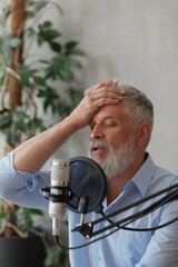 elderly man with gray hair is recording podcast in a recording studio with microphone and headphones. hand near face is gesture of forgetting and fatigue. senior radio host or interviewer.