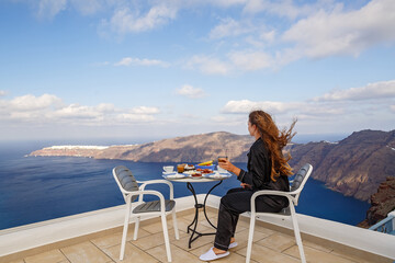 Woman is having breakfast at a table on a high terrace above the sea