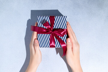 Female hands hold Blue and white stripe gift box with red bows on grey background. Copy space. Mockup. Flat lay.