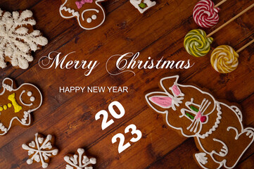 Merry Christmas and happy new year 2023 greeting concept - a brown wooden table background surrounded by various shaped gingerbreads with glaze and caramels. Top view.