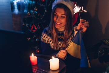 Celebrating Christmas and New year from distance online using laptop. Woman burns sparkler at home with Ukrainian flag
