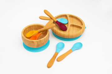 Six silicone spoons with handles of different colors and two bamboo plates with a suction cup on a white background. Cutlery is safe for babies