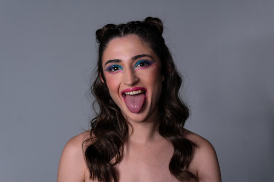 Portrait of funny young Caucasian woman having fun indoors, sticking out her tongue at camera. Close up of teenage girl faces at grey studio wall