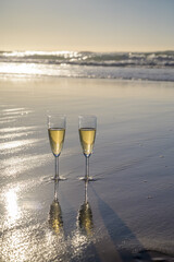 Glasses of cava or champagne sparkling wine on white sandy ocean beach with water waves on sunset in sunlights