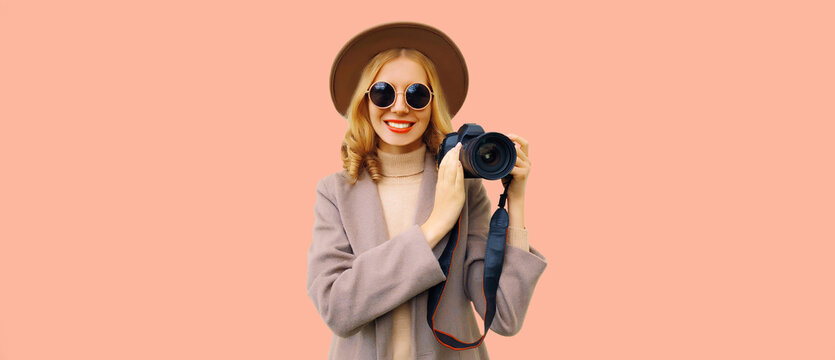 Portrait of stylish happy smiling woman photographer with digital camera taking picture wearing round hat, brown coat on background, blank copy space for advertising text