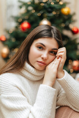 A girl with long hair in a knitted white sweater is sitting near the Christmas tree at home on Christmas. Close-up portrait of a girl.